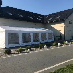 Biały namiot z przezroczystymi ścianami dołączony do budynku Domu Weselnego Olivia, otoczony dekoracyjnymi krzewami i kulami.