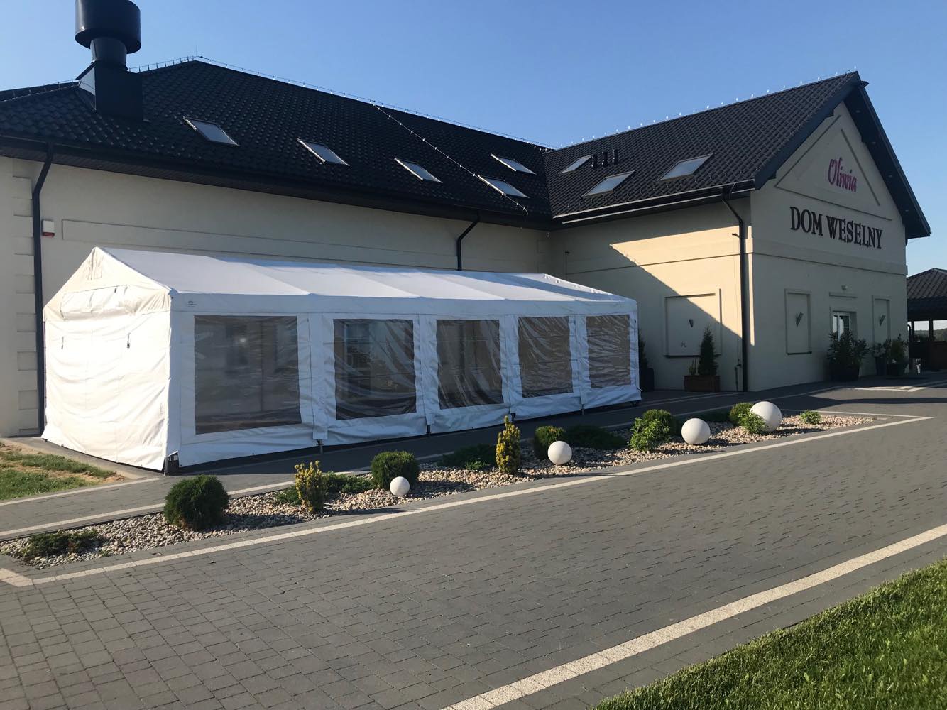 Biały namiot z przezroczystymi ścianami dołączony do budynku Domu Weselnego Olivia, otoczony dekoracyjnymi krzewami i kulami.