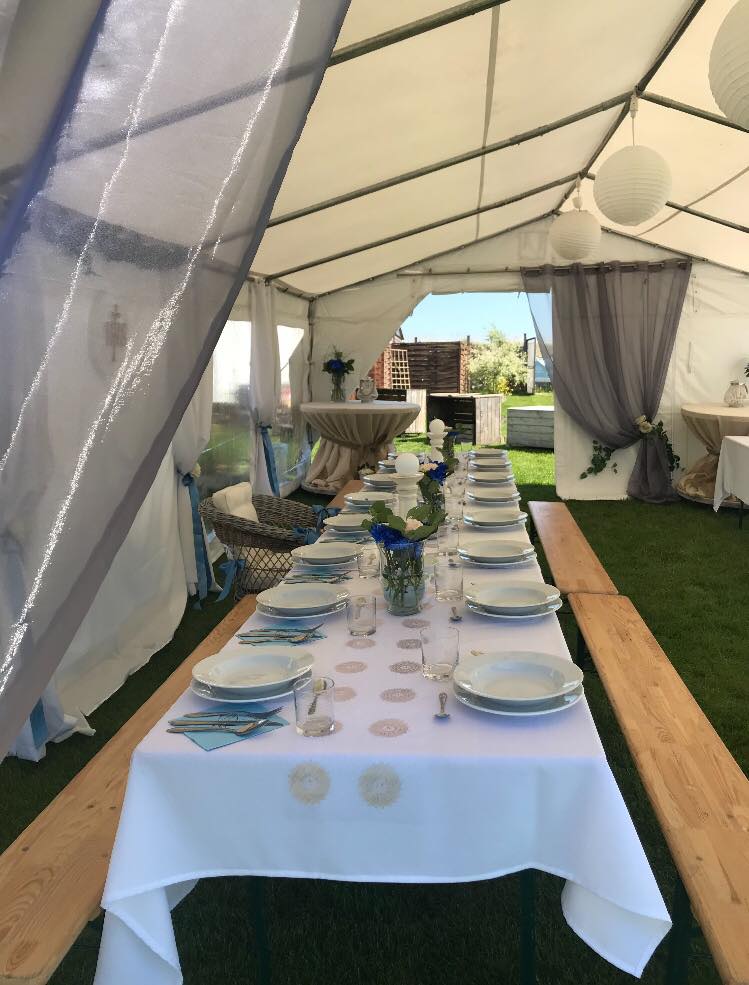 Uroczystość w białym namiocie z długim stołem nakrytym białym obrusem, zastawionym talerzami, sztućcami i szklankami, udekorowanym kwiatami w wazonach, z drewnianymi ławkami po bokach i papierowymi...