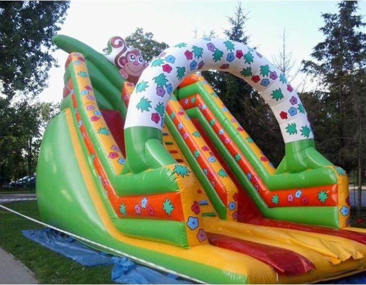 Zielony dmuchany zamek ze zjeżdżalnią, ozdobiony motywem małpki i kolorowymi kwiatami, ustawiony na trawie.