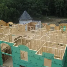 Wznoszenie konstrukcji domu o nietypowym kształcie z wieżyczką, widoczne drewniane szkielety ścian i zielona izolacja zewnętrzna, w tle las.