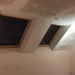 Prace wykończeniowe poddasza w Ełku: widoczne dwa okna dachowe w drewnianych ramach, osadzone w ścianie z płyt karton gips, sufit w trakcie szpachlowania.