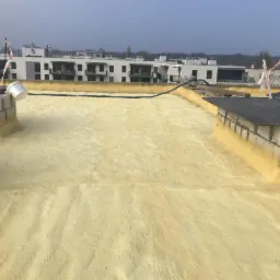 Żółta pianka PUR izolująca płaski dach budynku wielorodzinnego, widoczne kominy i elementy wentylacyjne oraz niebieski przewód. W tle zabudowa miejska.