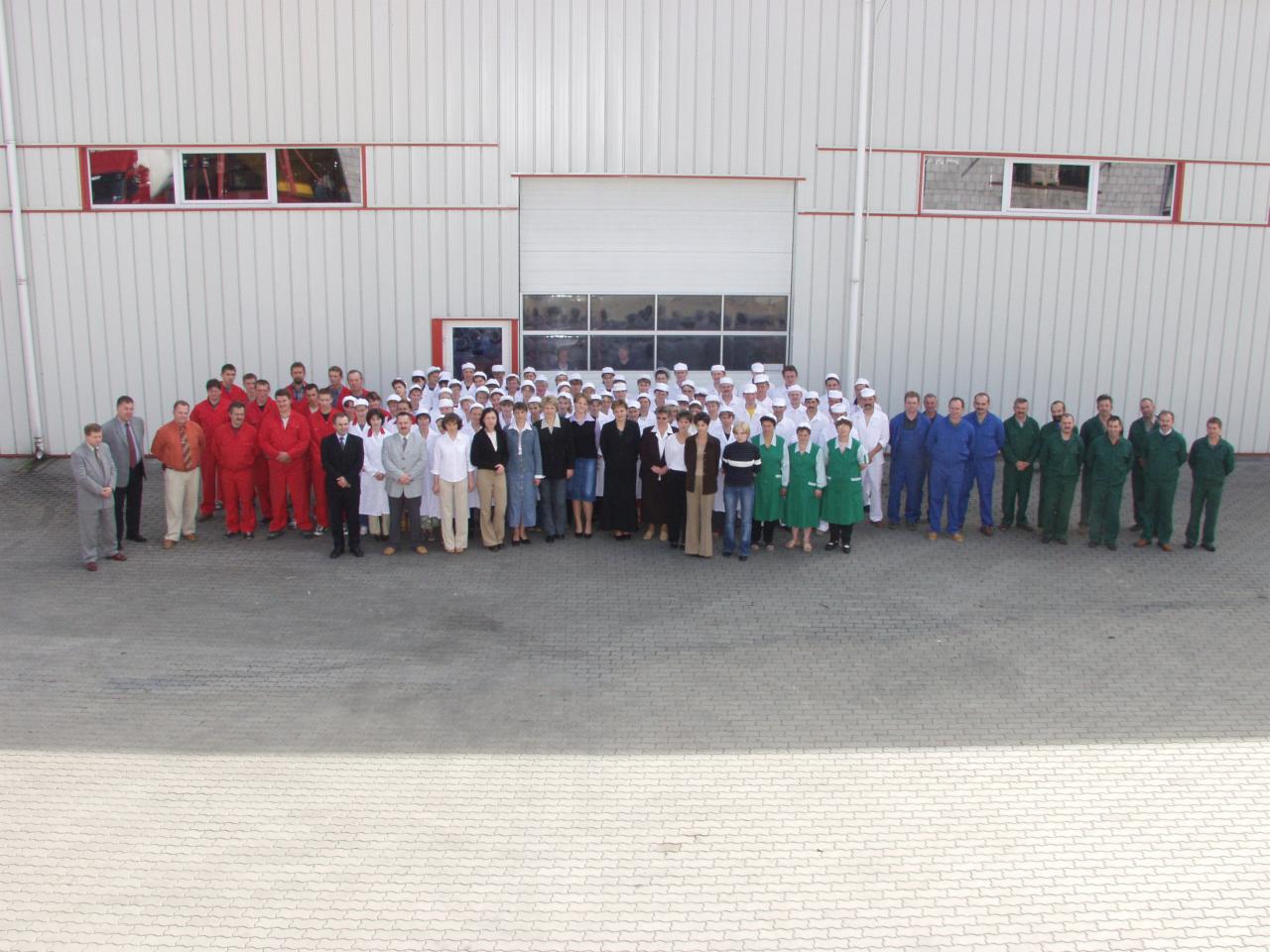 Duża grupa pracowników w różnorodnych strojach roboczych stoi przed budynkiem magazynowym z bramą rolowaną.