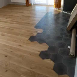 Połączenie jasnego parkietu z ciemnymi, heksagonalnymi płytkami podłogowymi w jasnym pomieszczeniu z grzejnikiem i zasłonami.