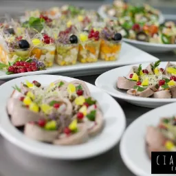 Elegancko podane przystawki: roladki mięsne dekorowane owocami i ziołami na białych talerzach oraz sałatki warstwowe w przezroczystych kubeczkach, całość na szarym blacie.