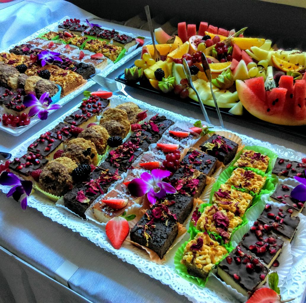 Dekoracyjnie ułożone ciasta, ciasteczka i owoce na dwóch paterach, ozdobione kwiatami, z widocznymi szczypcami do nakładania.