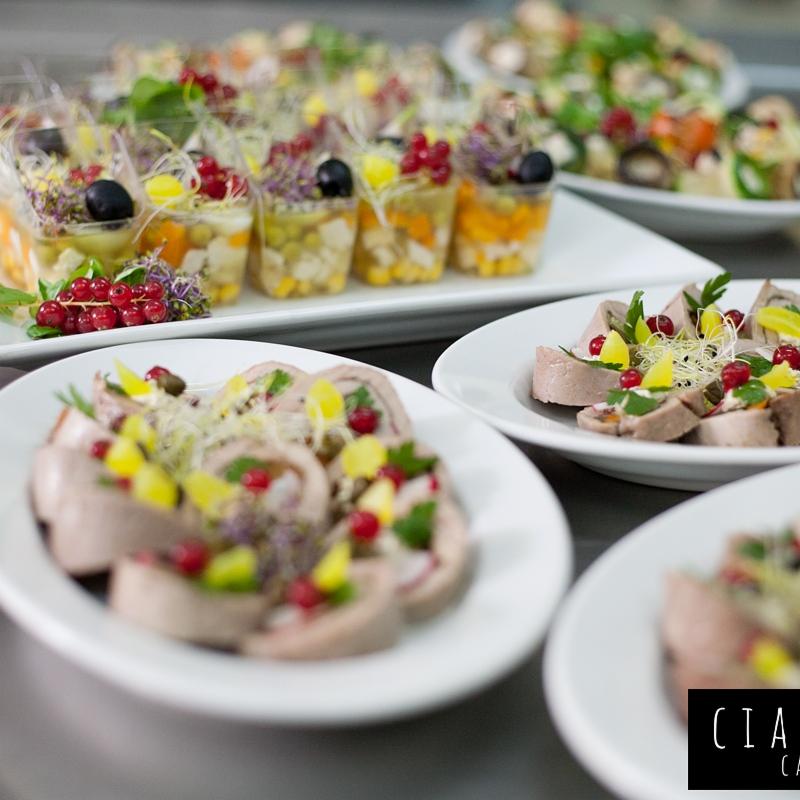 Elegancko podane przystawki: roladki mięsne dekorowane owocami i ziołami na białych talerzach oraz sałatki warstwowe w przezroczystych kubeczkach, całość na szarym blacie.