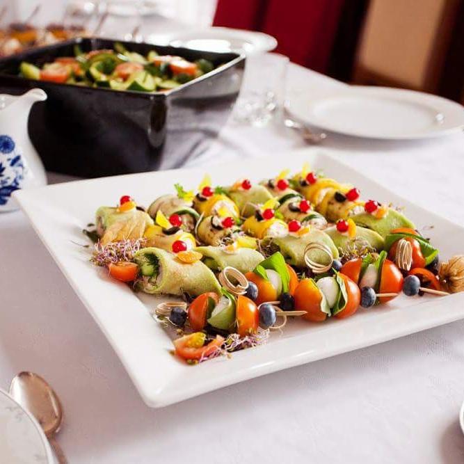 Elegancko podane przekąski na białym kwadratowym talerzu: roladki z cukinii z czerwonymi akcentami i szaszłyki z pomidorków koktajlowych, mozzarelli i borówek, w tle sałatka w czarnym naczyniu.