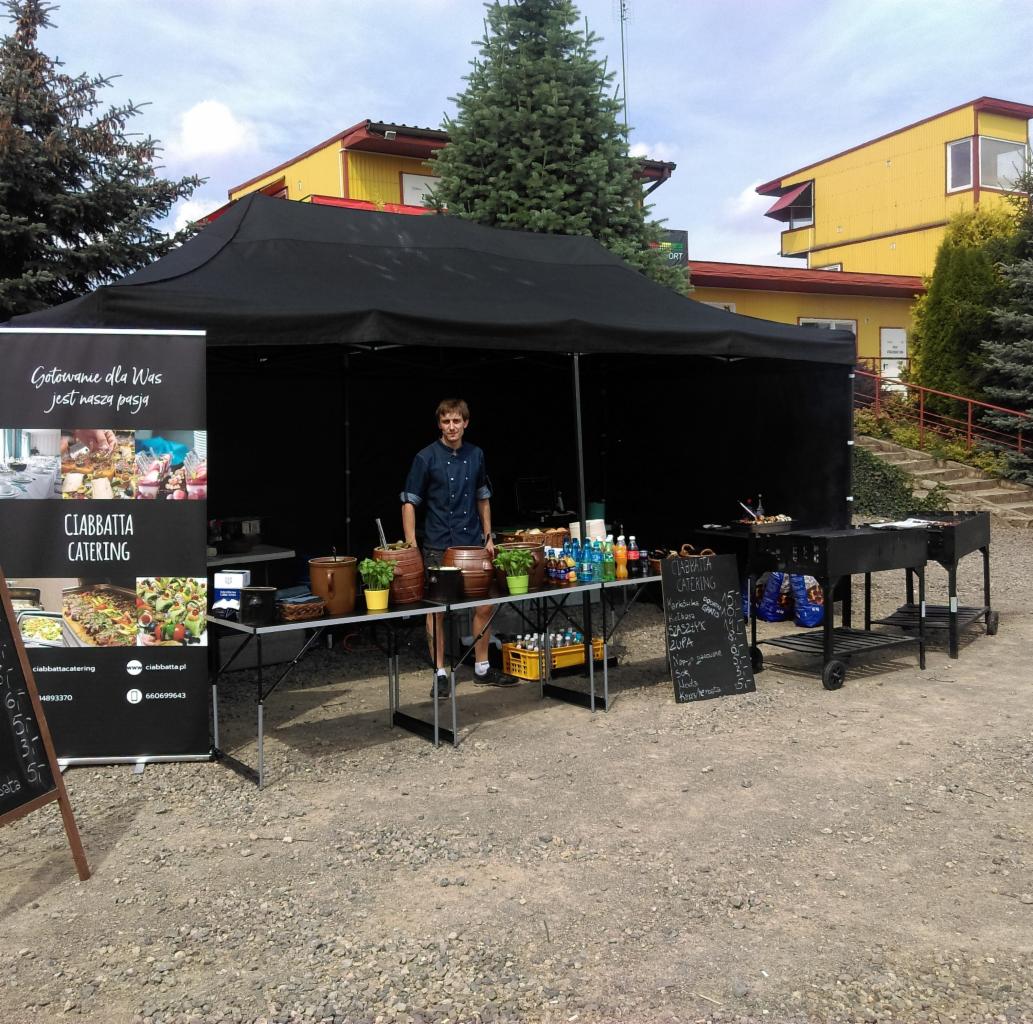 Stanowisko cateringowe pod czarnym namiotem z młodym mężczyzną w niebieskiej koszuli, menu wypisane kredą na tablicy, rośliny w doniczkach na stole, grill na kółkach w tle, reklama firmy...