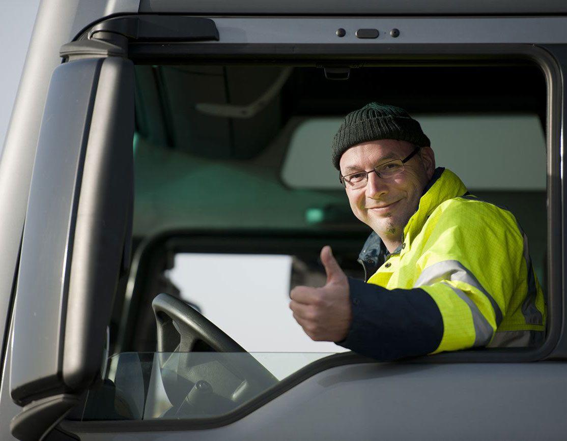 Uśmiechnięty kierowca w czapce i okularach, w żółtej kurtce odblaskowej, siedzący w kabinie ciężarówki i pokazujący kciuk w górę.