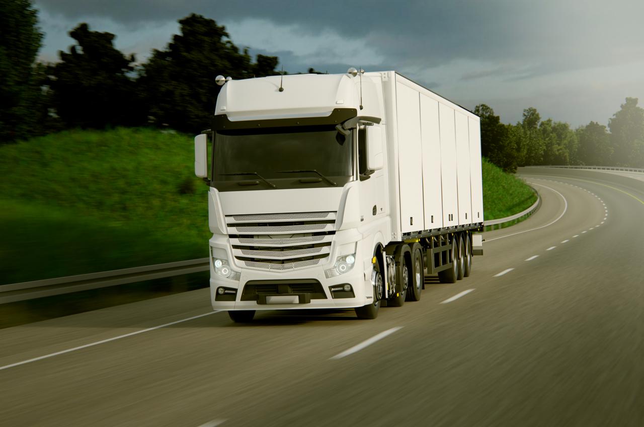 Biała ciężarówka z naczepą, widok z przodu, jadąca po asfaltowej drodze w słoneczny dzień, z zielonymi wzgórzami i drzewami w tle.