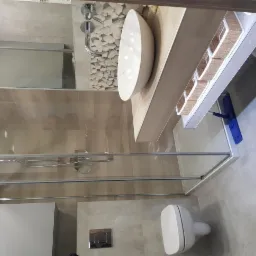 Nowoczesna łazienka z kabiną prysznicową ze szklanymi drzwiami, umywalką nablatową w kształcie elipsy, wiszącą toaletą i mopem do podłogi opartym o półkę.