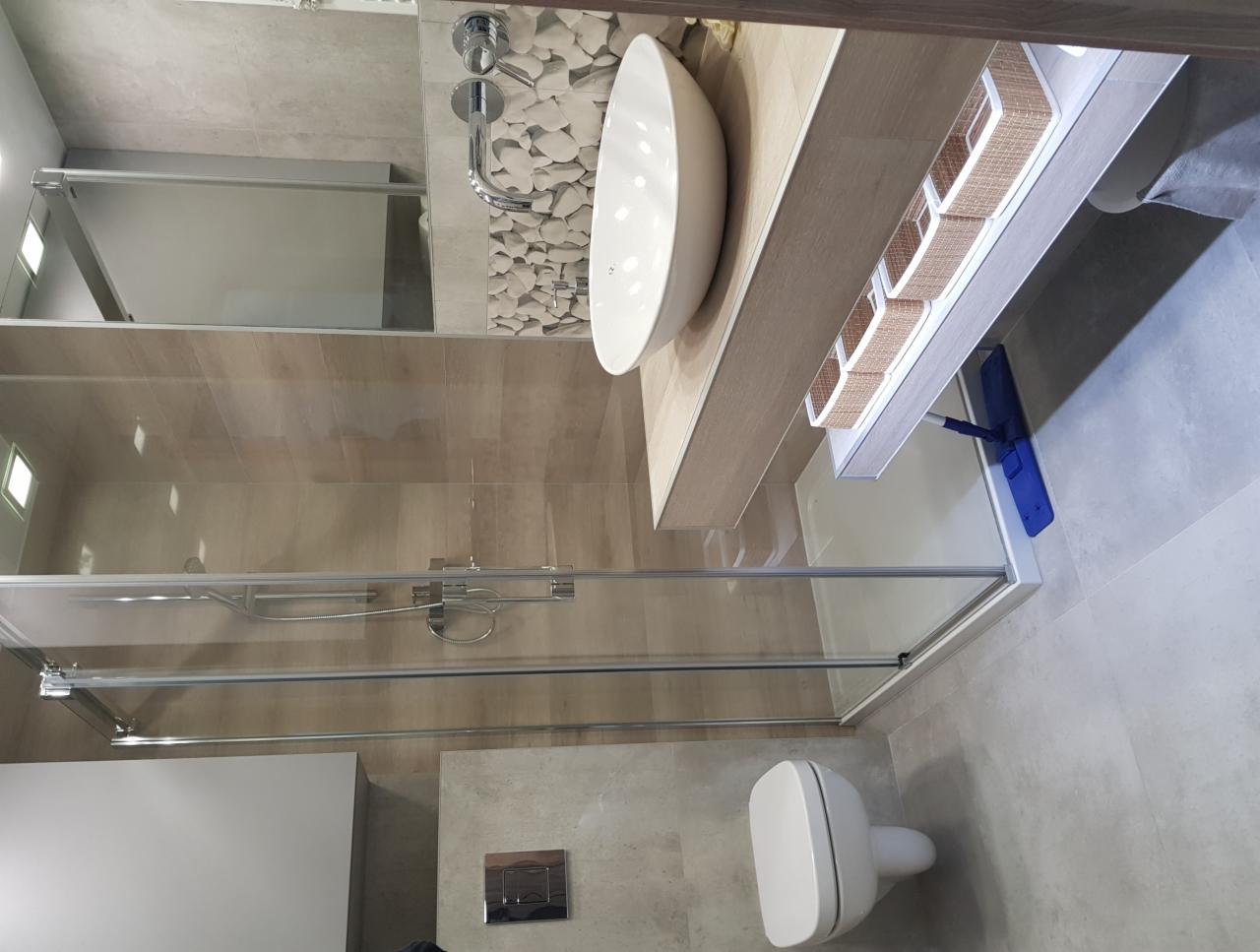 Nowoczesna łazienka z kabiną prysznicową ze szklanymi drzwiami, umywalką nablatową w kształcie elipsy, wiszącą toaletą i mopem do podłogi opartym o półkę.