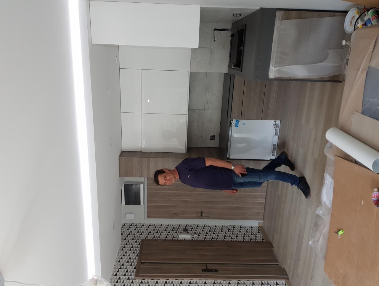 Nowoczesna kuchnia w trakcie montażu: białe szafki, drewnopodobne panele, czarne detale, widoczny pracownik w jeansach na tle częściowo zabezpieczonej podłogi.