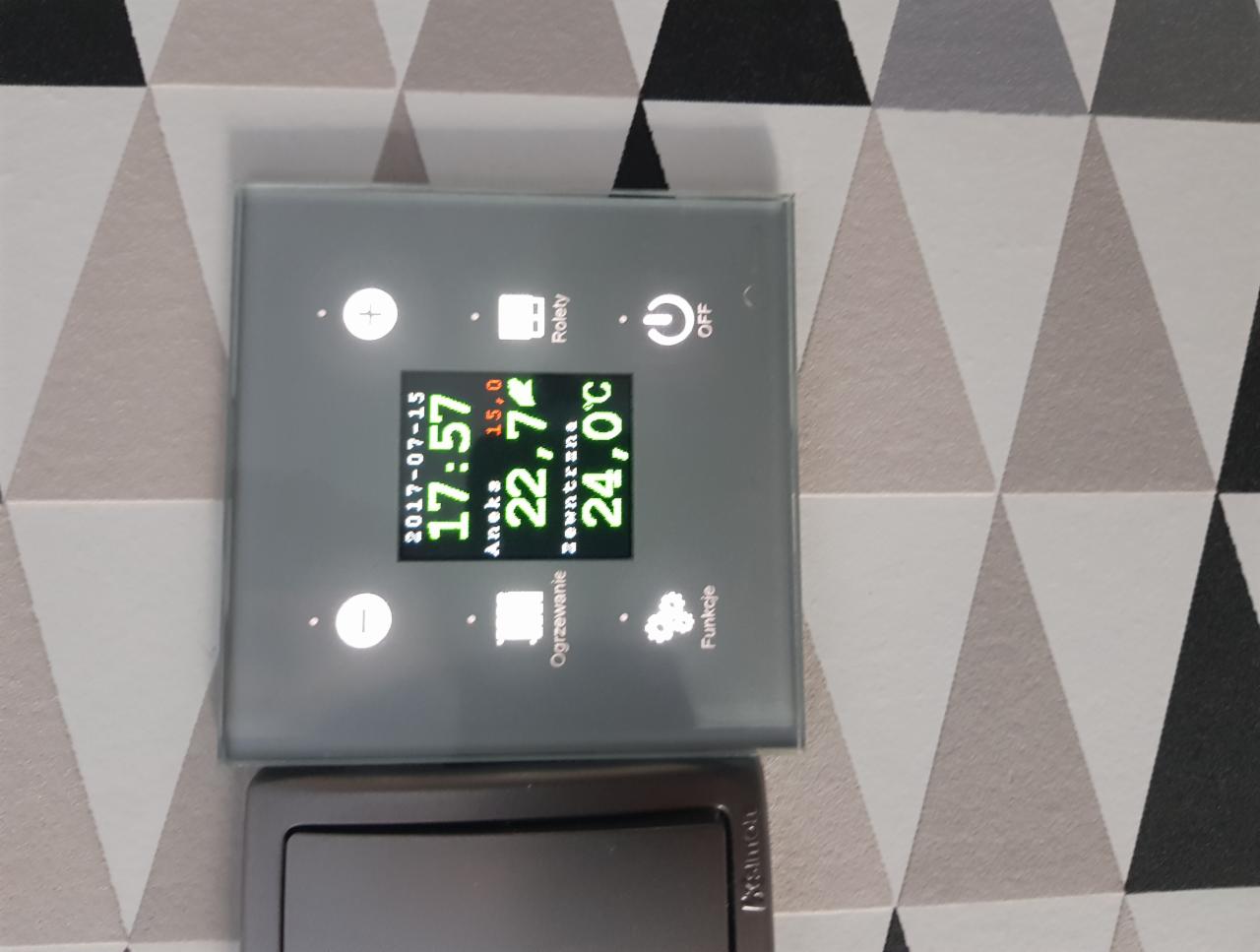 Szklany panel sterowania inteligentnym domem z wyświetlaczem pokazującym datę, godzinę i temperaturę wewnętrzną oraz zewnętrzną, zamontowany na wzorzystej ścianie.