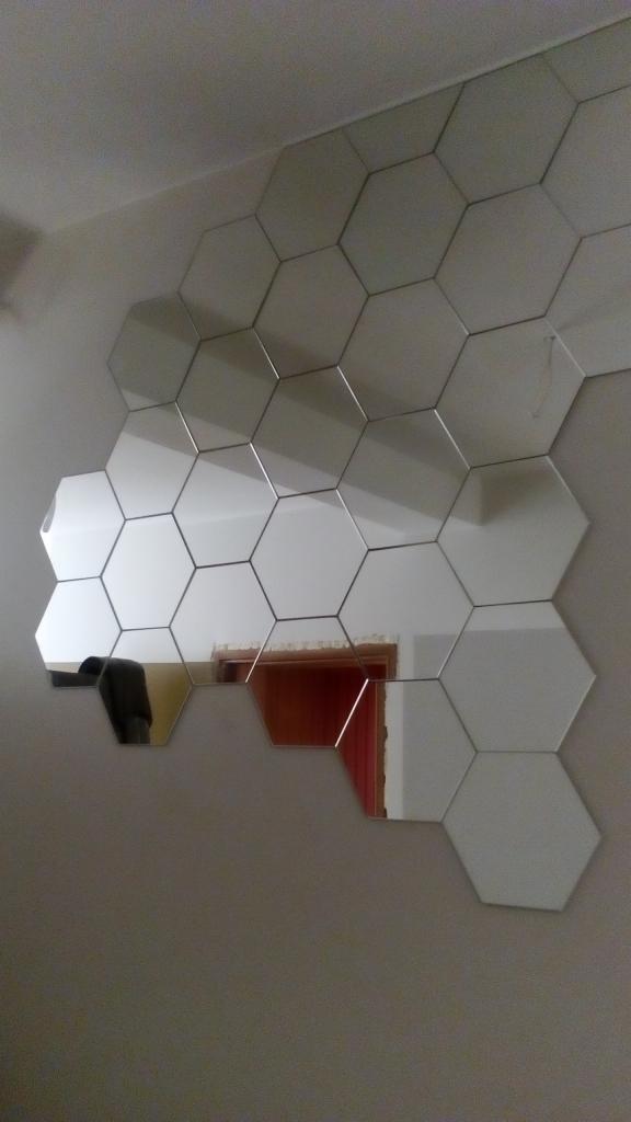 Dekoracyjna ściana z heksagonalnych luster, odbijająca fragment wnętrza z czerwonymi drzwiami i ciemnym meblem.