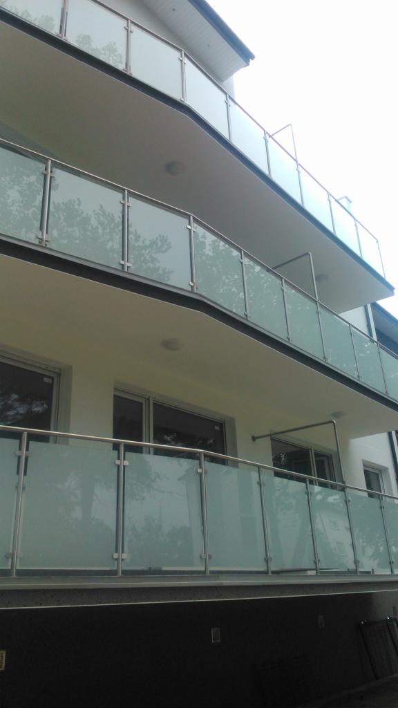 Szklane balustrady balkonowe z metalowymi elementami mocującymi, widok z dołu na trzy kondygnacje, odbicie drzew na szkle.