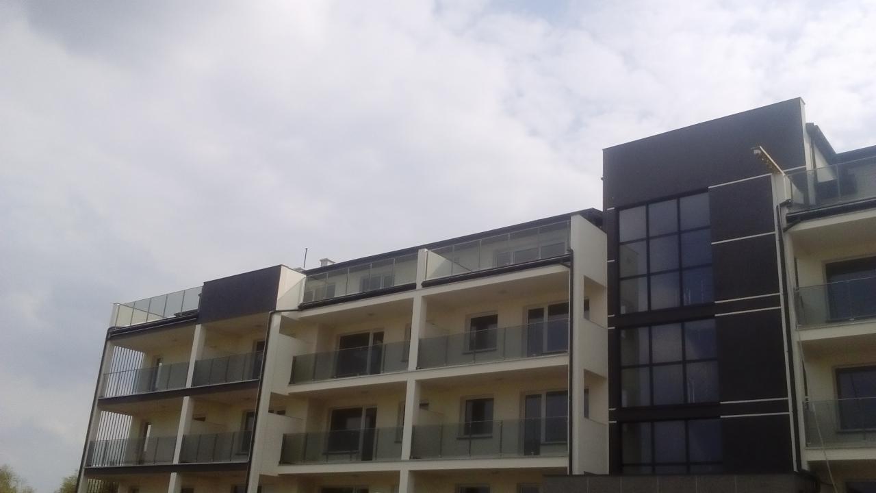 Nowoczesny budynek mieszkalny z balkonami i szklanymi balustradami, widok z dołu na tle pochmurnego nieba.