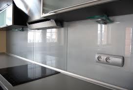 Biała szklana ściana nad blatem kuchennym z widocznym gniazdkiem elektrycznym i fragmentem okapu.