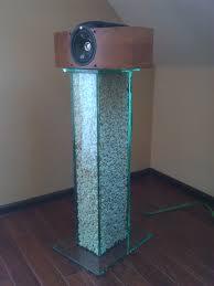 Szklana kolumna ekspozycyjna z wypełnieniem z drobnych kamyczków, zwieńczona drewnianą obudową z głośnikiem. Widoczna podstawa i górny element kolumny wykonane ze szkła.