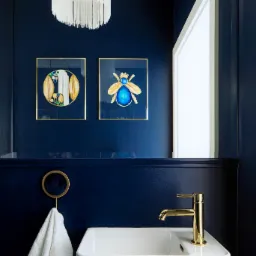 Elegancka łazienka w odcieniach granatu z białymi elementami: umywalka, ręcznik z frędzlami i żyrandol z wiszącymi nitkami. Na ścianie wiszą złote ramy z abstrakcyjnymi obrazami owadów.