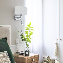 MiluMila - Jasna sypialnia z drewnianą szafką nocną, na której stoi szklany wazon z zielonymi gałązkami, świeca zapachowa, książki, obok łóżko z zielonymi poduszkami, na ścianie biała lampa