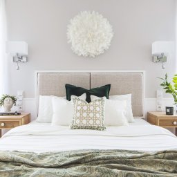 MiluMila - Jasna sypialnia z beżowym zagłówkiem, białymi zasłonami i dekoracyjną, okrągłą ozdobą z piór nad łóżkiem. Na szafkach nocnych lampki i dekoracje roślinne, a na łóżku poduszki w odcieniach bieli...