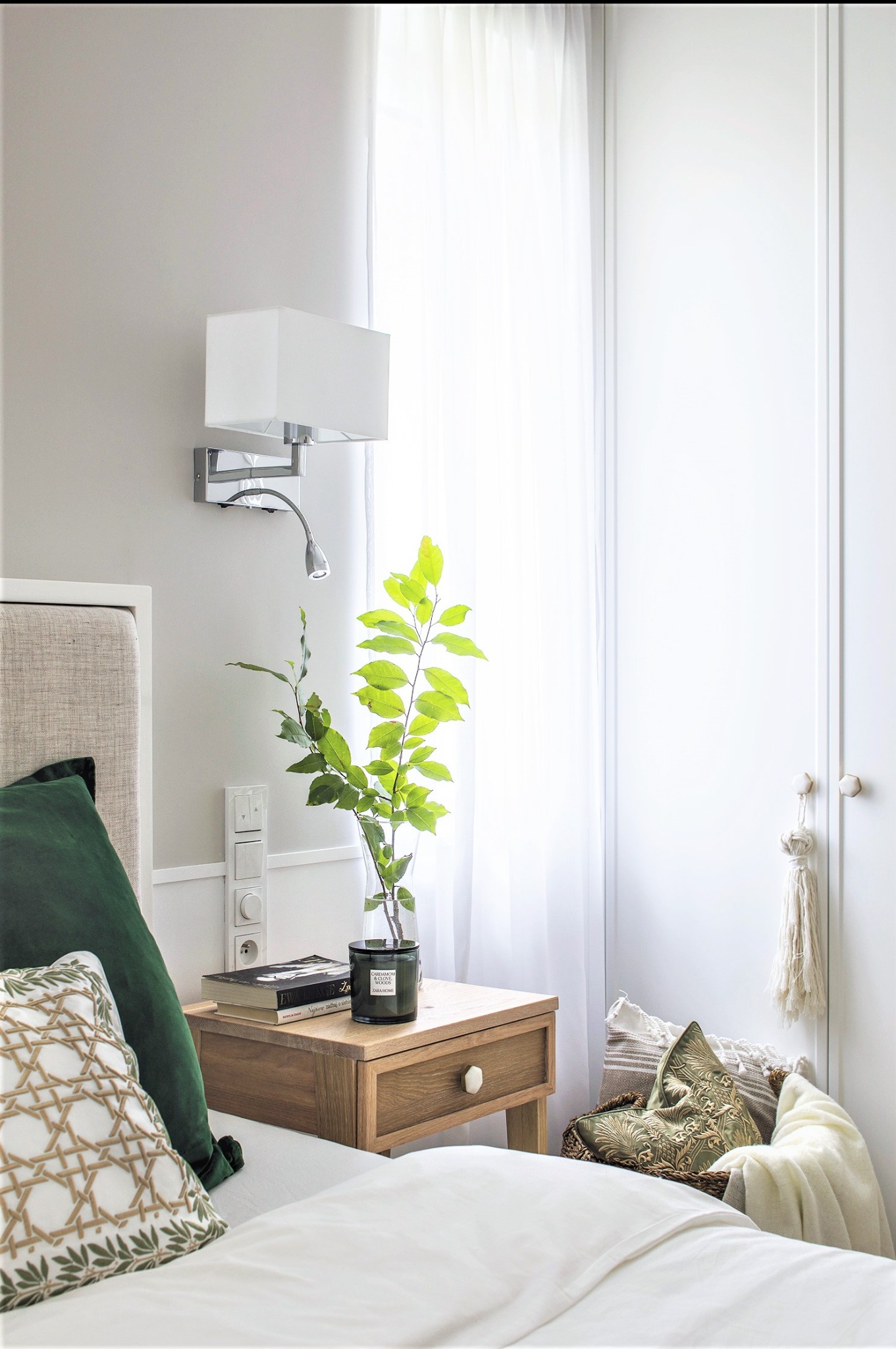 Jasna sypialnia z drewnianą szafką nocną, na której stoi szklany wazon z zielonymi gałązkami, świeca zapachowa, książki, obok łóżko z zielonymi poduszkami, na ścianie biała lampa