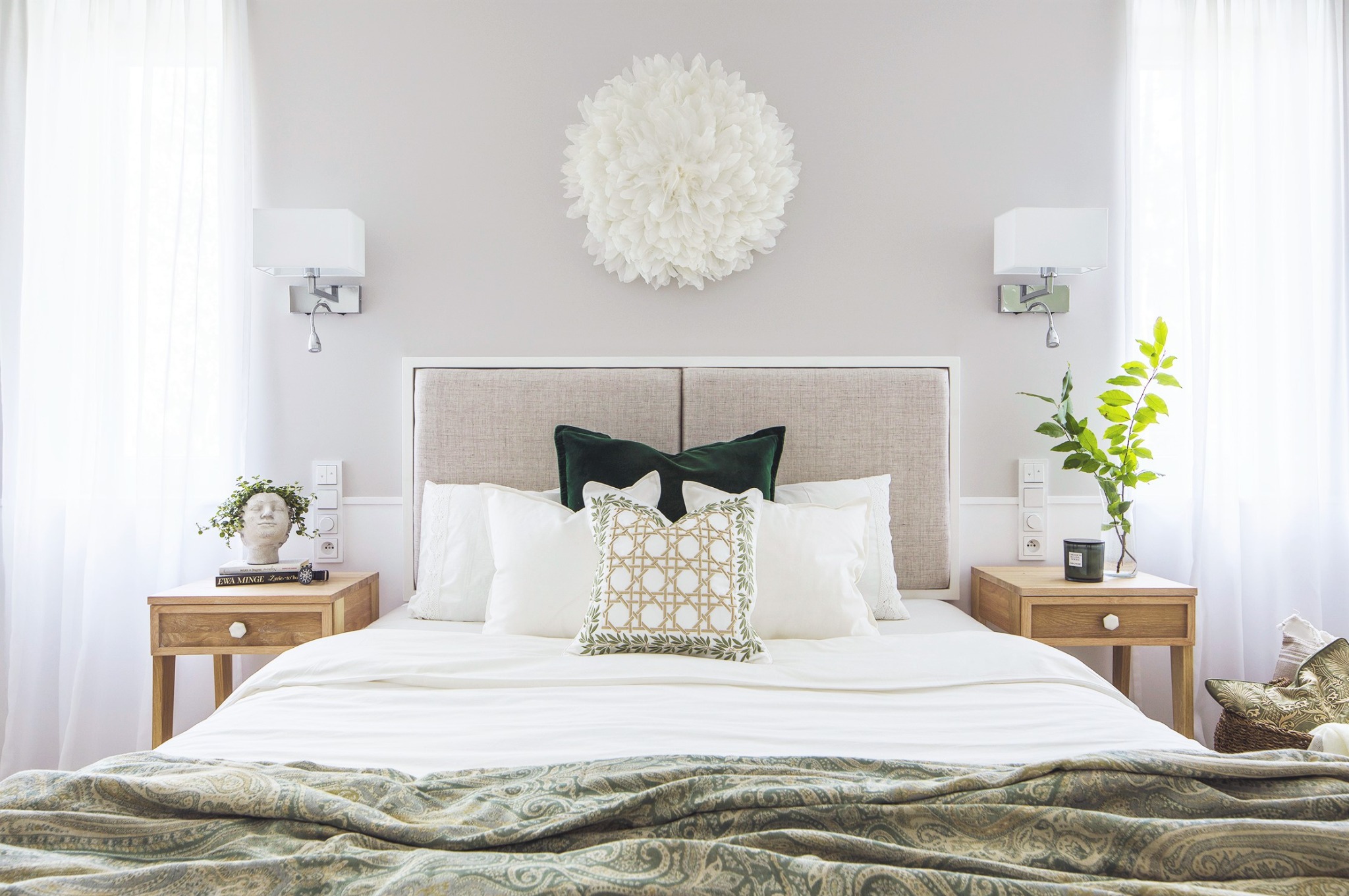 Jasna sypialnia z beżowym zagłówkiem, białymi zasłonami i dekoracyjną, okrągłą ozdobą z piór nad łóżkiem. Na szafkach nocnych lampki i dekoracje roślinne, a na łóżku poduszki w odcieniach bieli...