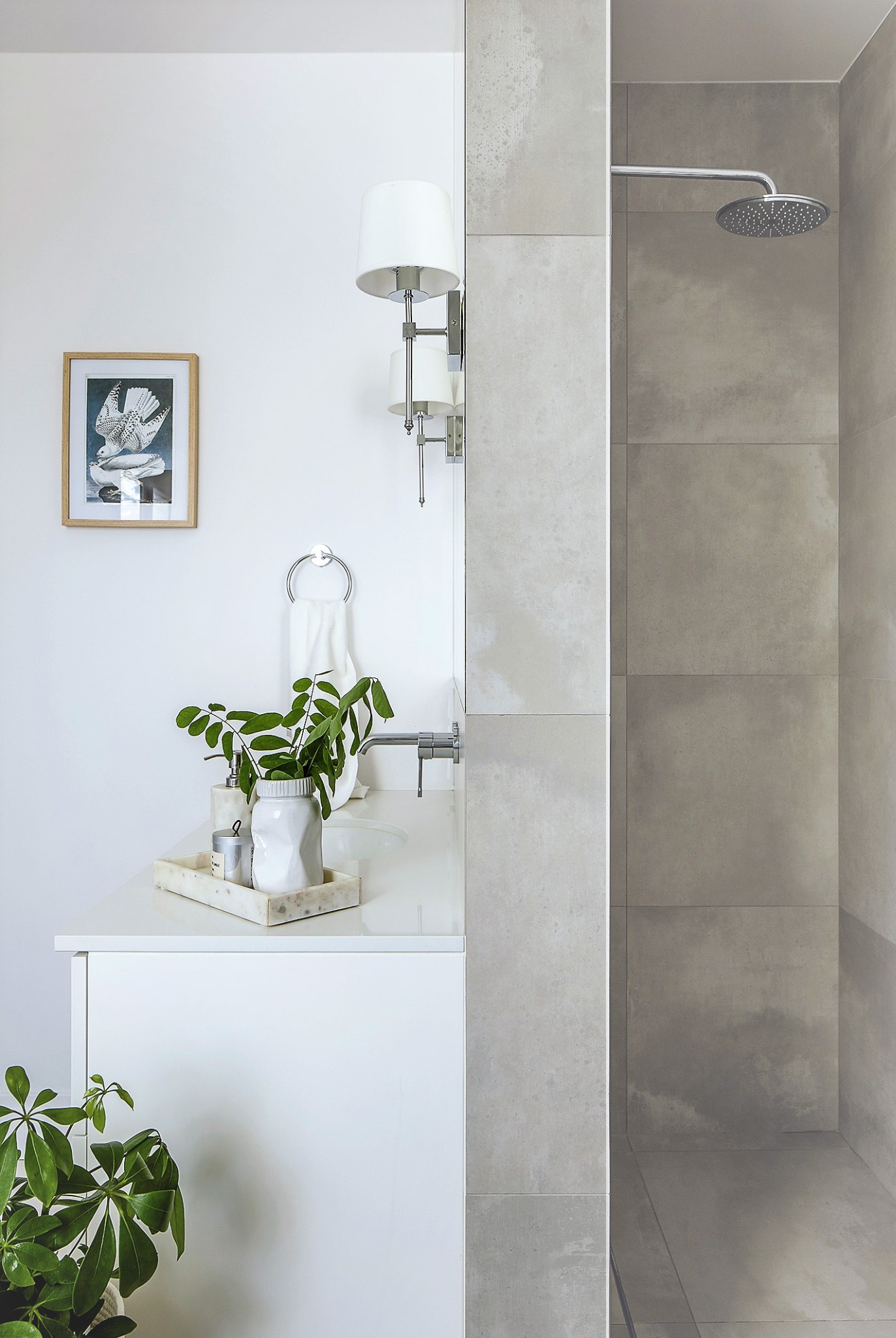 Nowoczesna łazienka z białą szafką pod umywalką, marmurową tacą z akcesoriami, roślinami w wazonie i deszczownicą w kabinie prysznicowej wyłożonej szarymi płytkami.