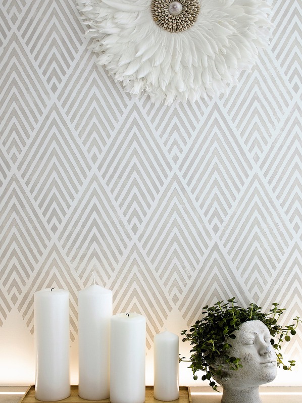 Dekoracyjna ściana z geometrycznym wzorem w odcieniach beżu, ozdobiona białym pióropuszem z muszelkowym środkiem, obok zestaw białych świec różnej wysokości i głowa z roślinami.