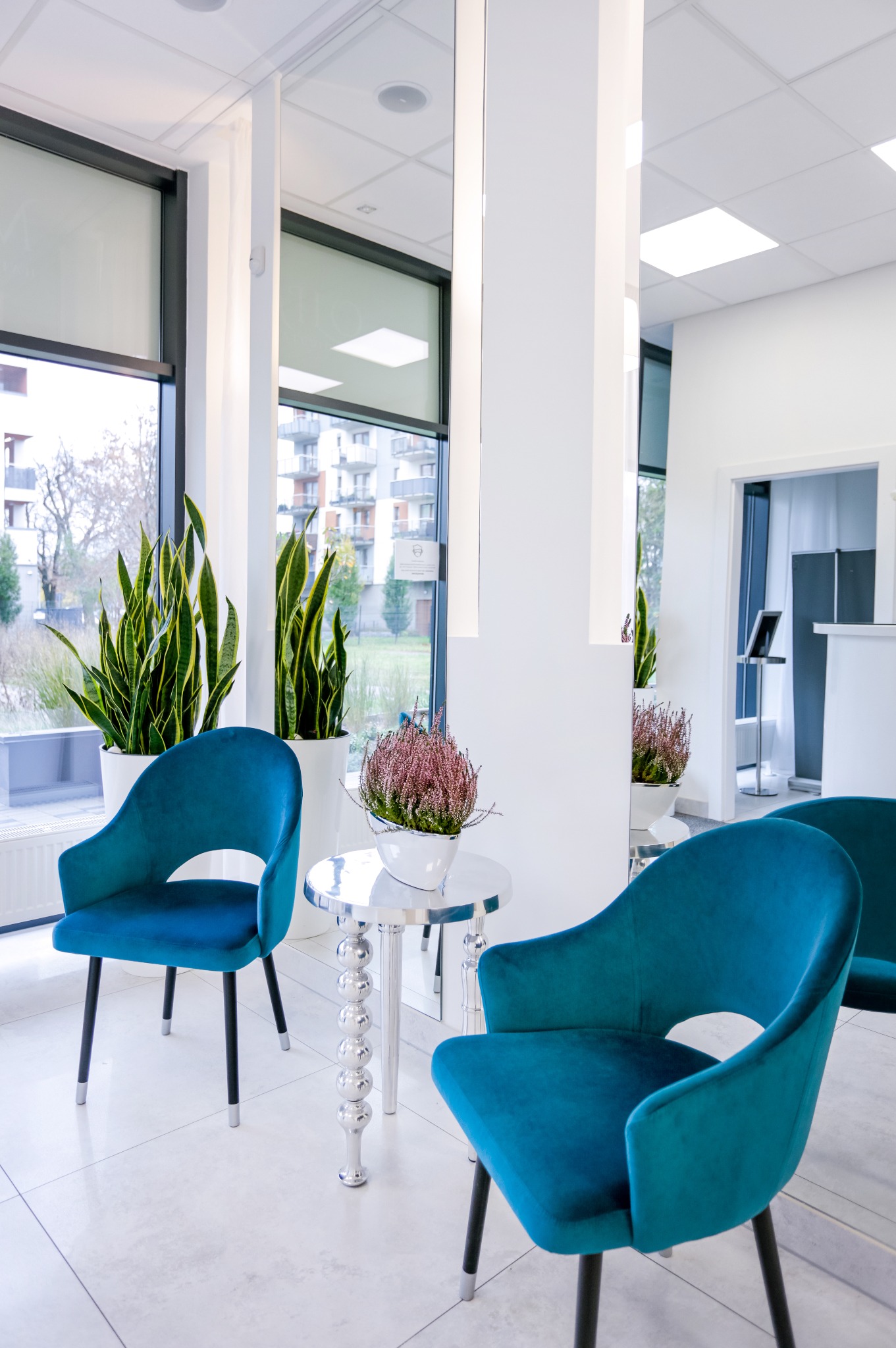 Jasne wnętrze biura z dwoma turkusowymi krzesłami, okrągłym stolikiem z metalową nogą i roślinami doniczkowymi przy oknie.