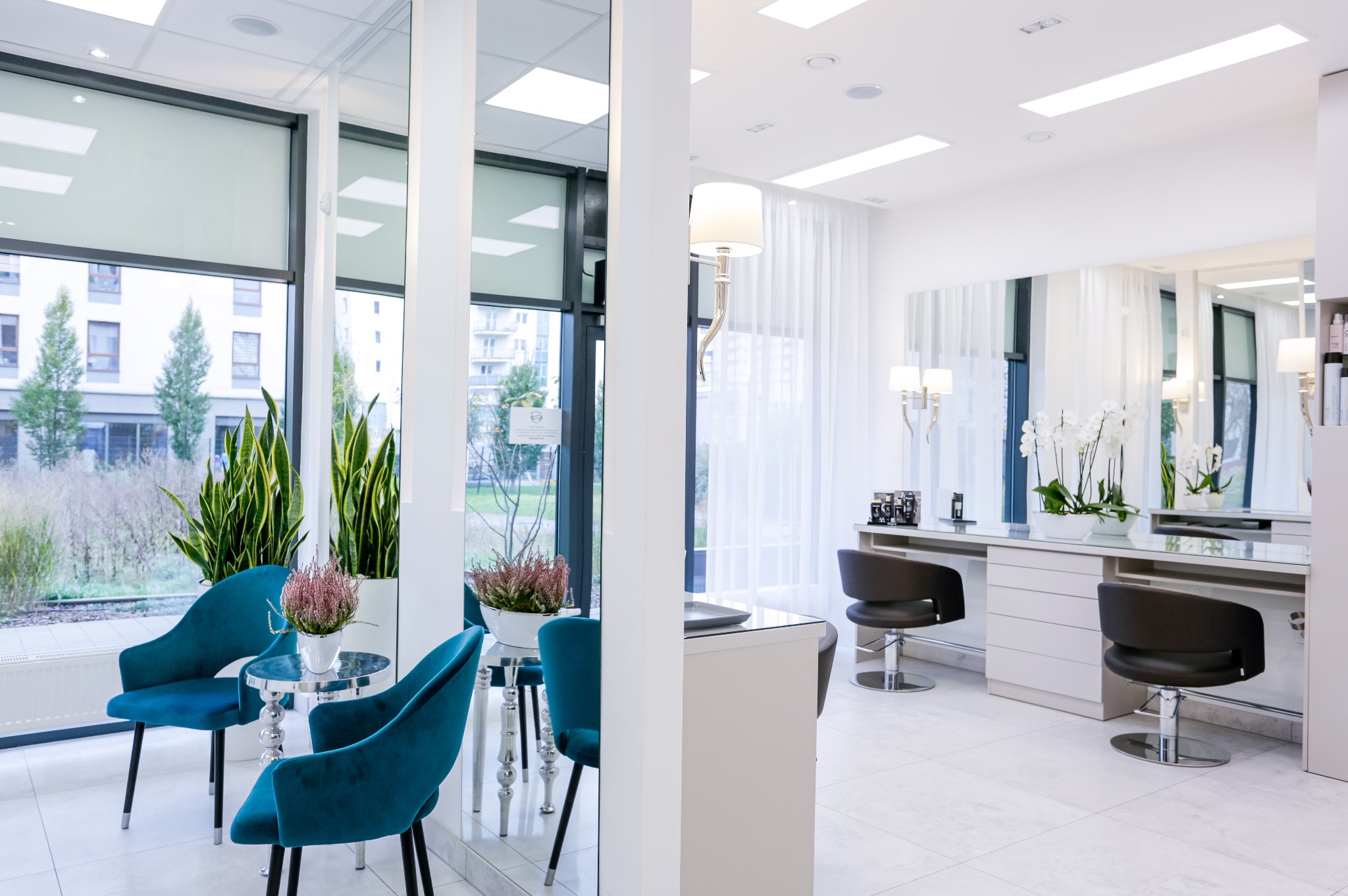 Jasne wnętrze salonu kosmetycznego z turkusowymi fotelami dla klientów i stanowiskami do manicure z lustrami, widok przez okno na budynek mieszkalny.