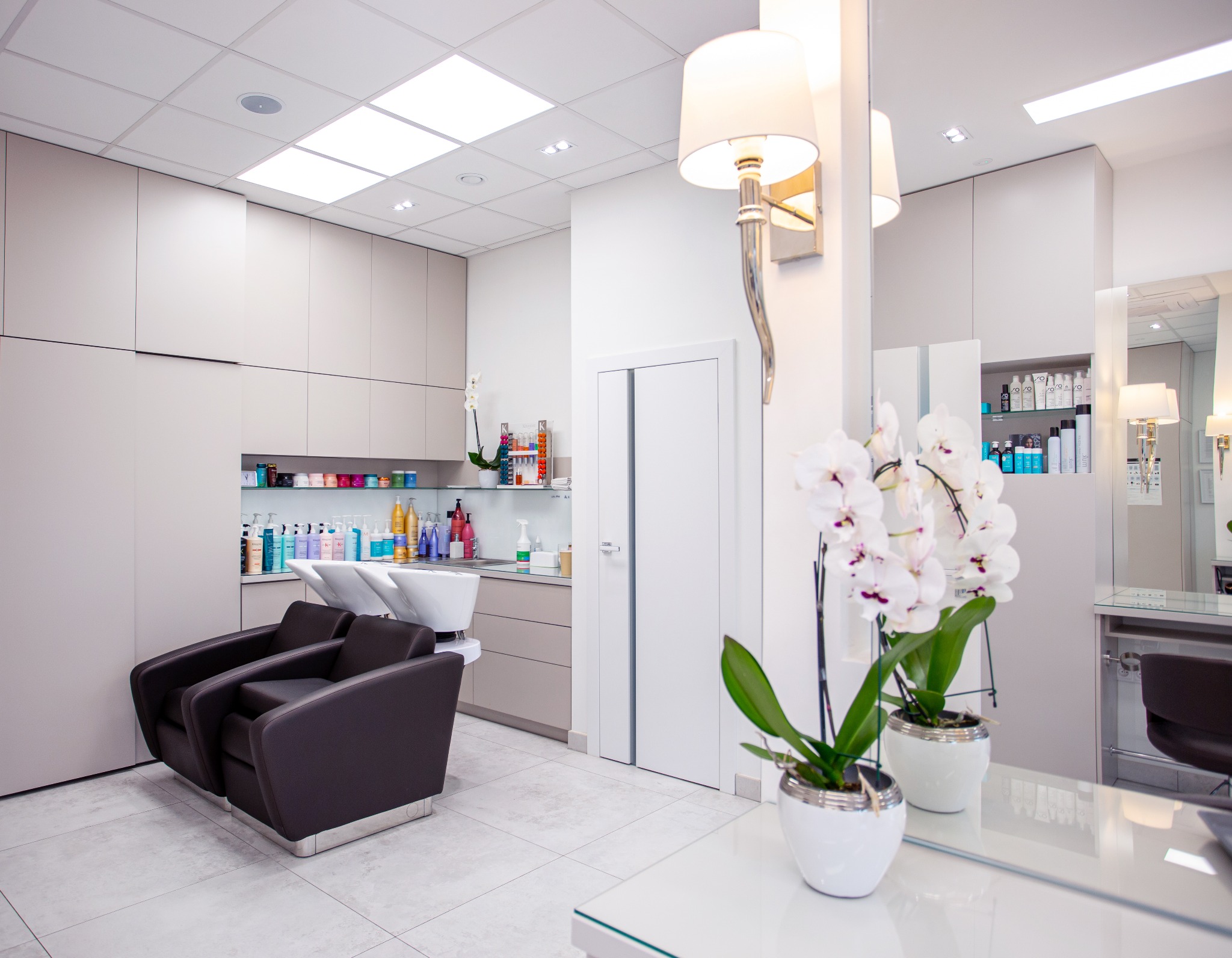 Nowoczesne wnętrze salonu fryzjerskiego z dwoma fotelami, stanowiskami do mycia włosów, półkami z kosmetykami, lustrem i dekoracyjnymi lampami ściennymi, oświetlone białym światłem.