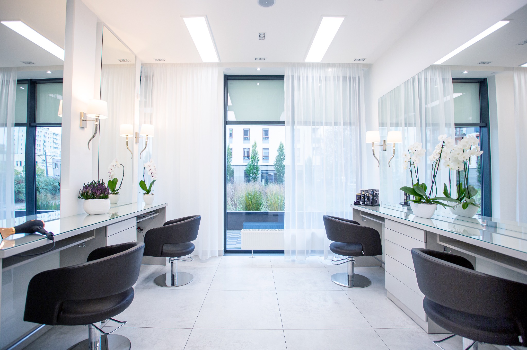 Jasne, przestronne wnętrze salonu fryzjerskiego z dwoma stanowiskami, duże okno z widokiem na zieleń, białe zasłony i płytki podłogowe, czarne krzesła obrotowe, lustra z oświetleniem i kwiaty...