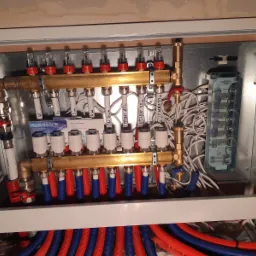 Rozdzielacz ogrzewania podłogowego w skrzynce, widoczne rury czerwone i niebieskie, zawory, przepływomierze oraz okablowanie elektryczne, z bliska.