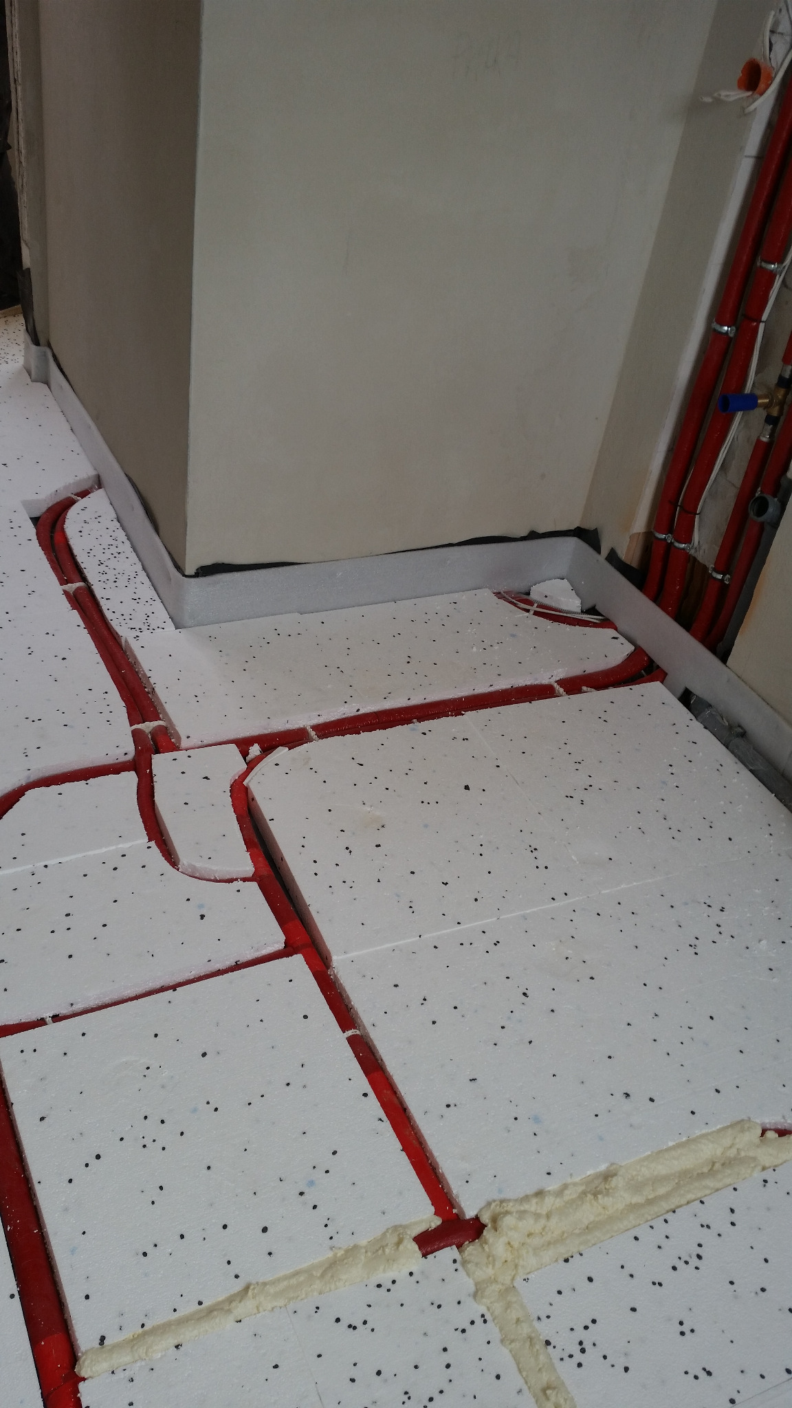 Przygotowanie do wylewki pod ogrzewanie podłogowe: czerwone rury w wycięciach styropianu, szczeliny wypełnione pianką montażową, widoczne fragmenty instalacji grzewczej przy ścianie.