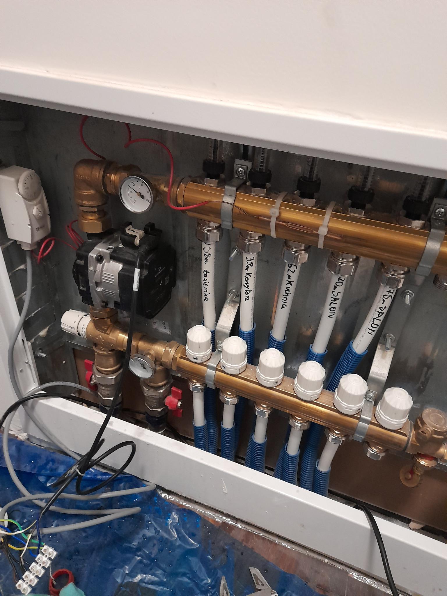 Rozdzielacz ogrzewania podłogowego z rurami oznaczonymi nazwami pomieszczeń (korytarz, kuchnia, salon, łazienka), widoczna pompa obiegowa i manometr, instalacja w trakcie montażu, zabezpieczona folią.