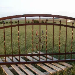 Brązowe, metalowe przęsło ogrodzeniowe z dekoracyjnymi elementami w kształcie liści i skręconych prętów, leżące na drewnianej palecie na tle zielonego trawnika i drzew