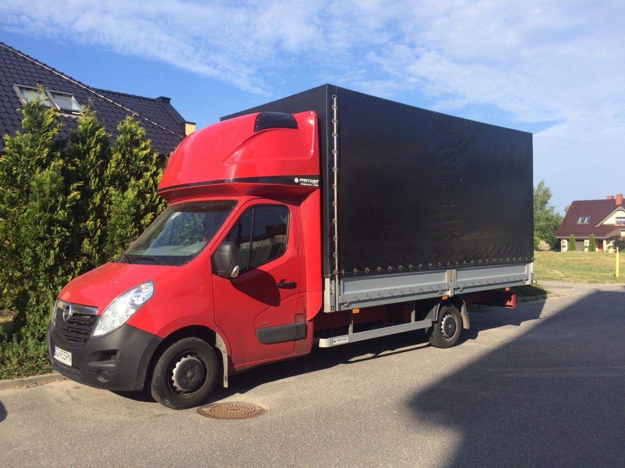 Czerwony samochód dostawczy z czarną plandeką zaparkowany na ulicy, widoczny z boku na tle domów i zieleni.