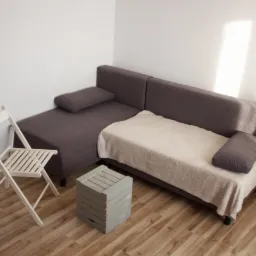 Szary narożnik z beżowym pledem, białe składane krzesło i szary stolik nocny z drewnianych skrzynek na parkiecie.