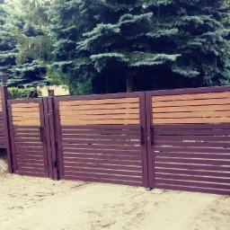 Nowoczesne ogrodzenie posesji z brązowych, poziomych profili metalowych i wstawkami z drewna kompozytowego, na tle zielonych drzew iglastych.