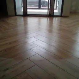 Ułożona podłoga z paneli w jodełkę, widoczne ślady po kleju lub lakierze, drzwi balkonowe w tle.