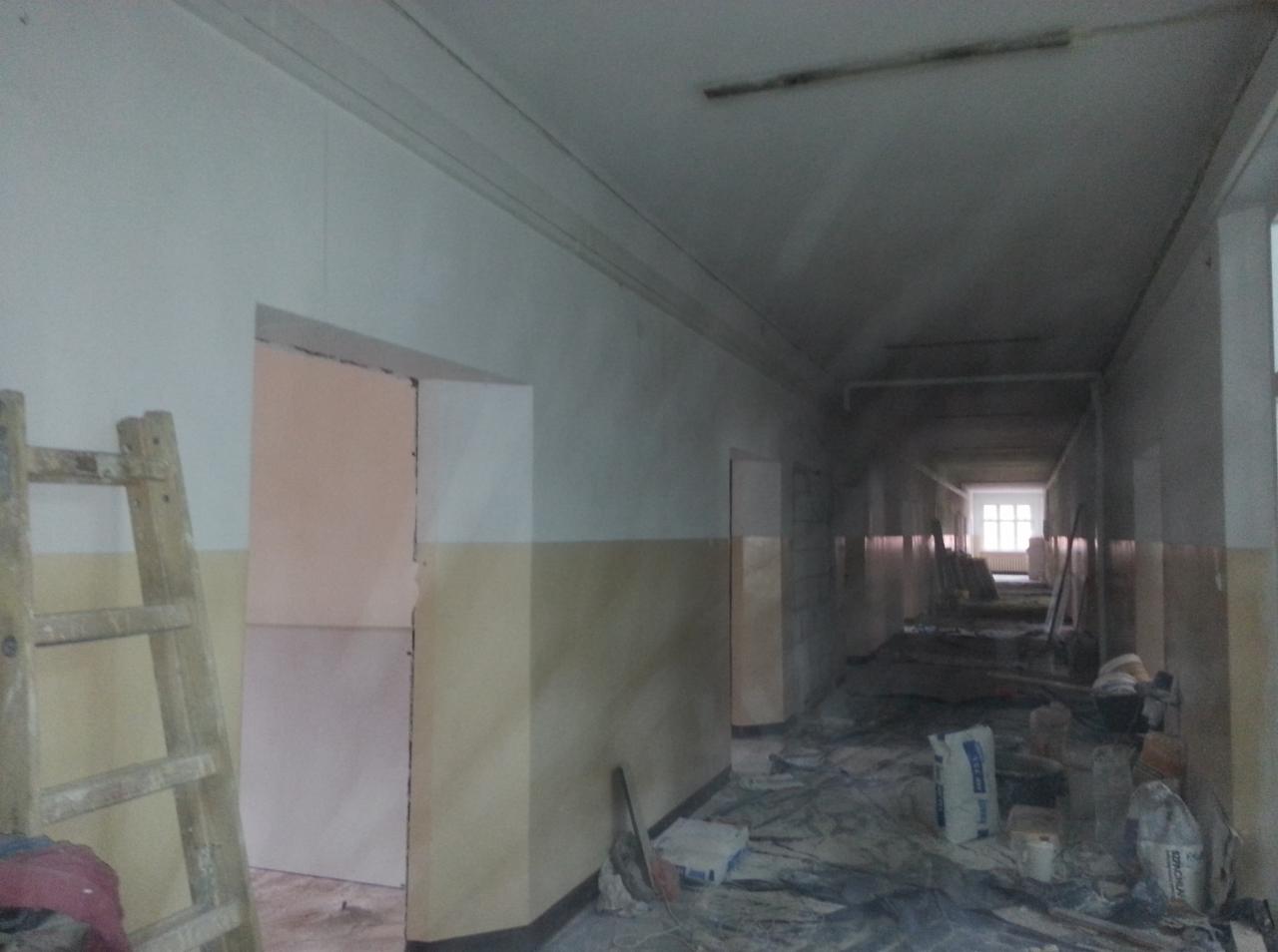 Korytarz w trakcie remontu z widoczną drabiną, świeżo pomalowanymi ścianami do połowy na beżowo, workami z zaprawą i folią ochronną na podłodze.