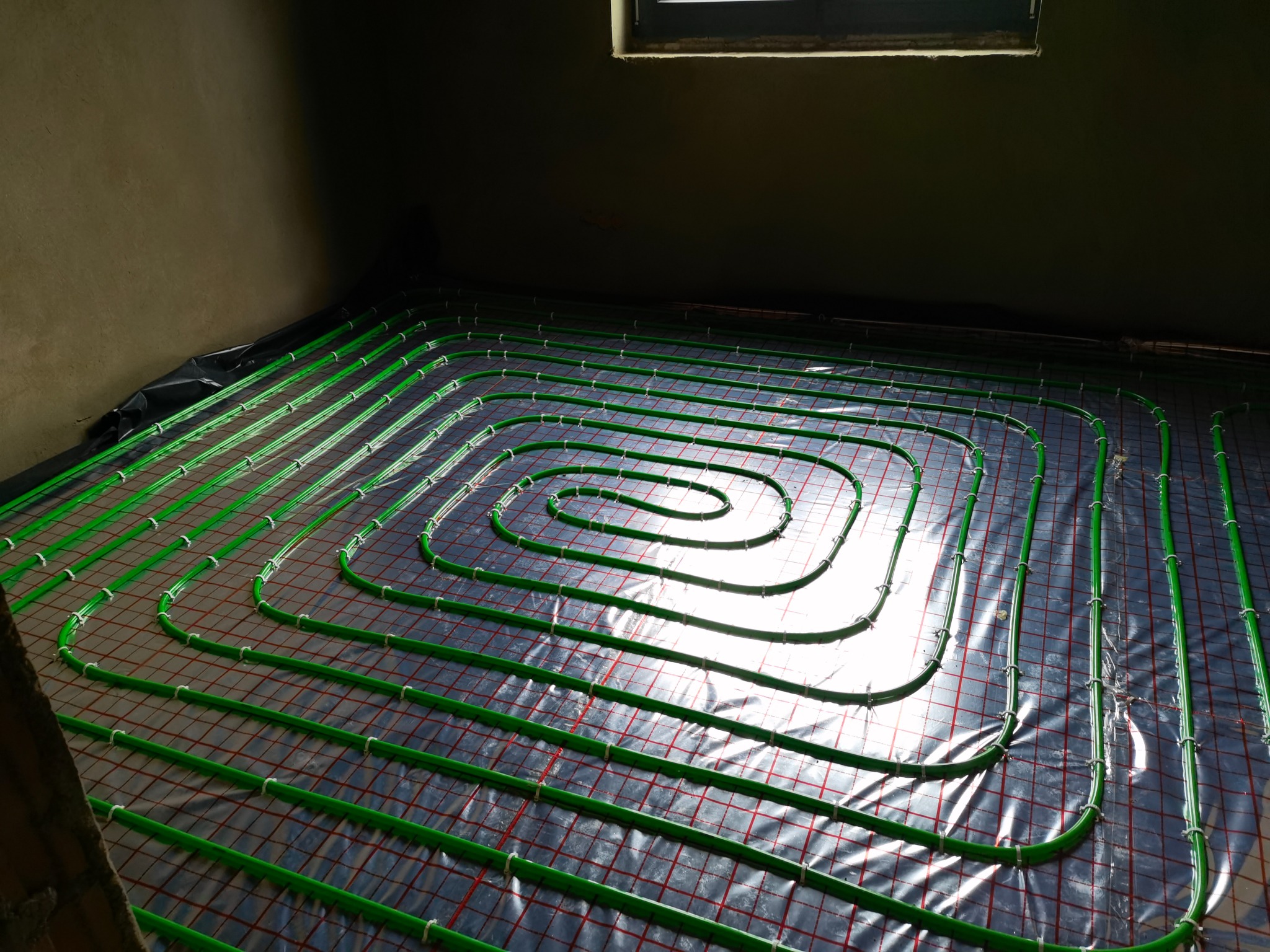 Świeżo ułożone, spiralne rury ogrzewania podłogowego w kolorze zielonym, przymocowane do siatki montażowej, pokryte folią izolacyjną, widoczne w surowym wnętrzu z oknem u góry.