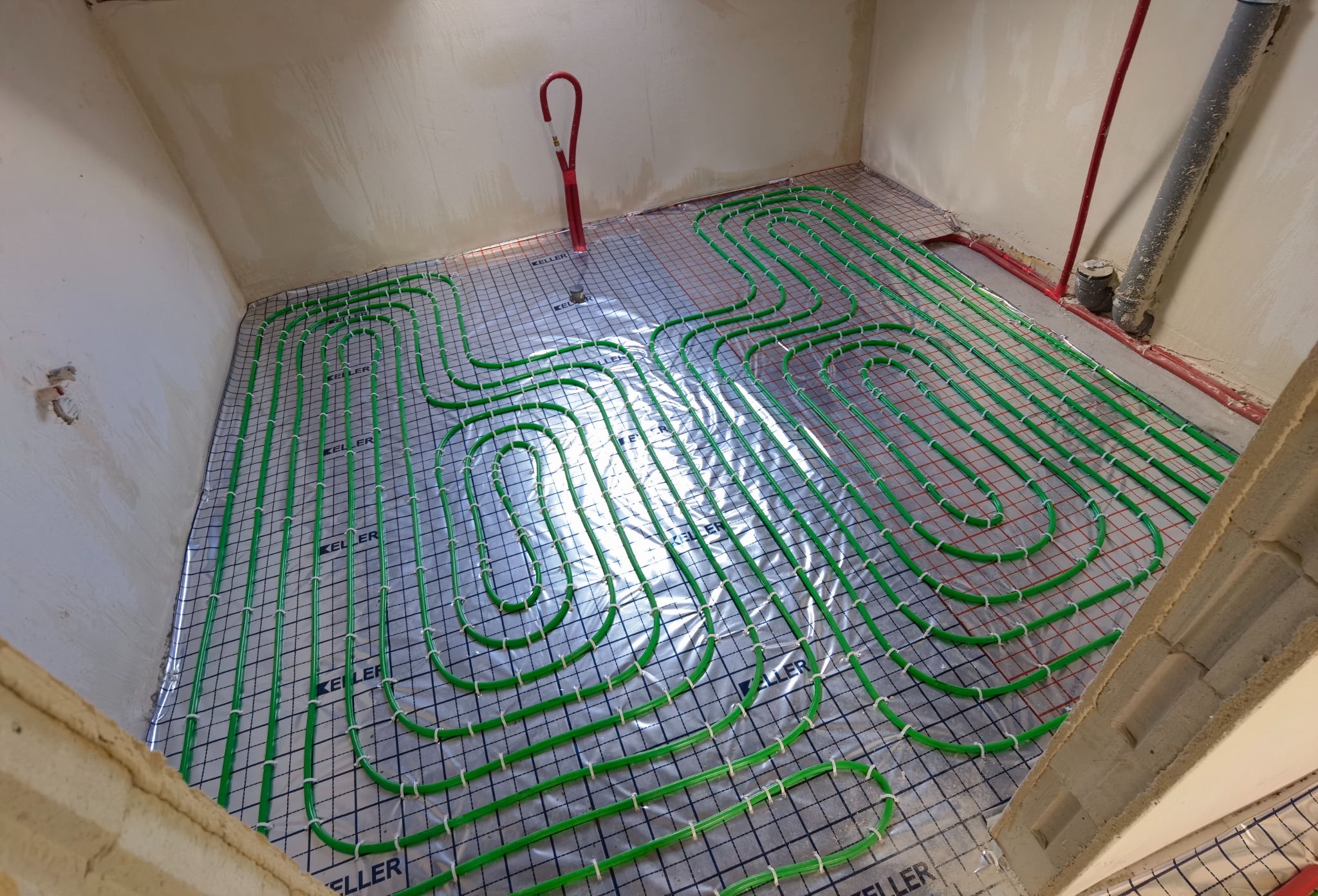 Montaż zielonych rur ogrzewania podłogowego na srebrnej macie z siatką w surowym pomieszczeniu, widoczne fragmenty ścian i pionowe rury.