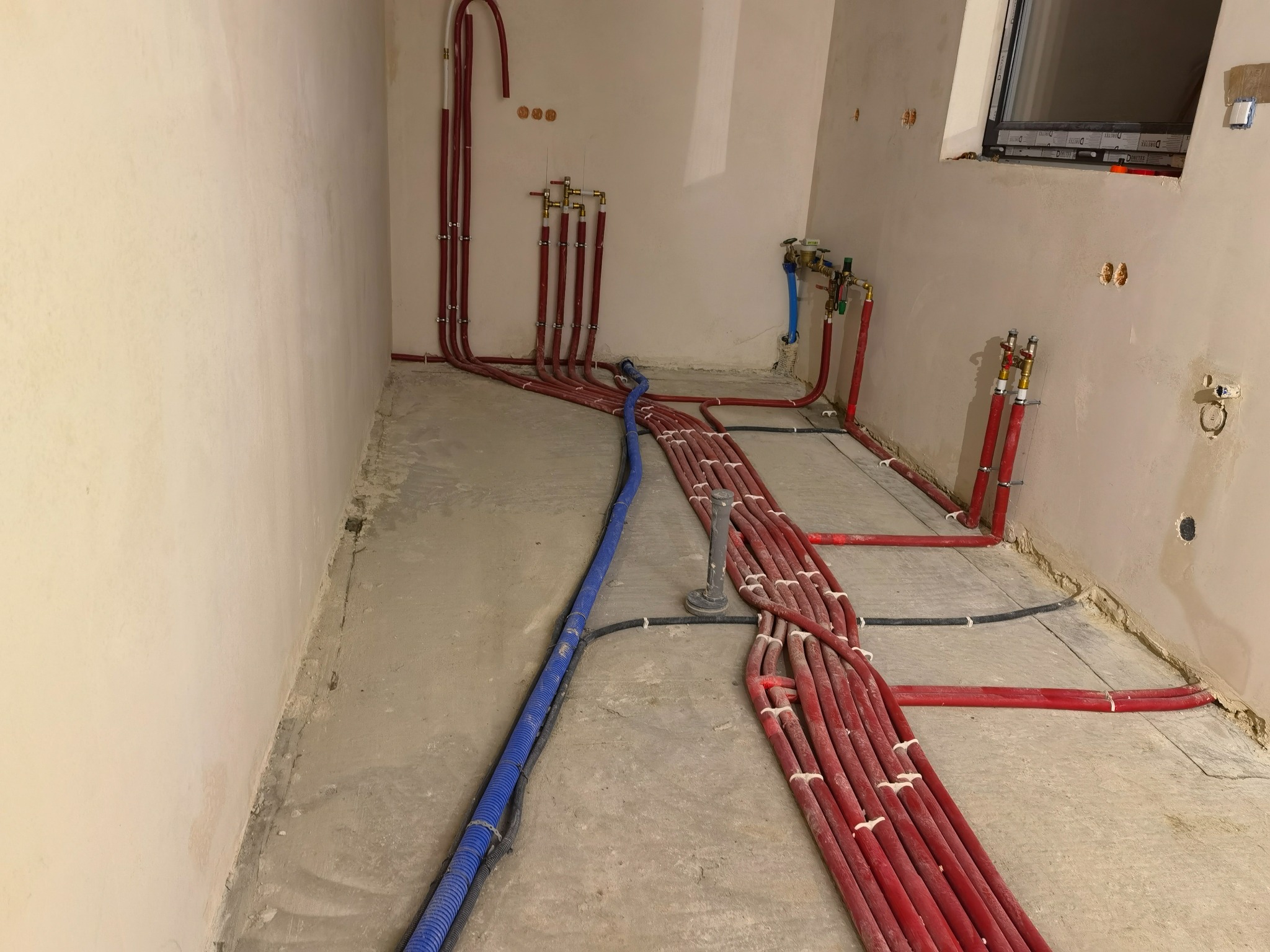 Czerwone i niebieskie rury instalacji grzewczej/hydraulicznej ułożone na surowym betonie w pomieszczeniu w trakcie remontu, widoczne kolektory i przyłącza.