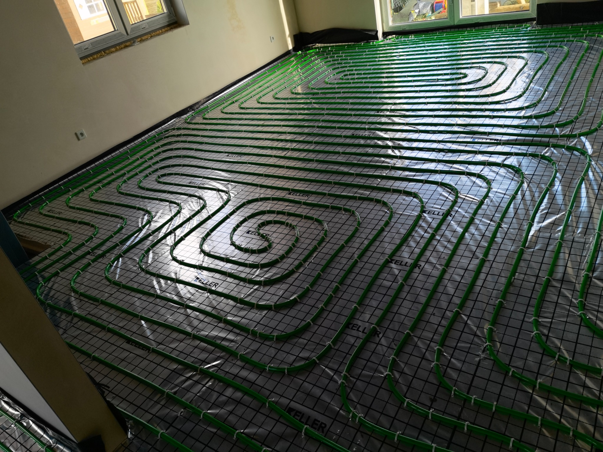 Zielone rury ogrzewania podłogowego ułożone na folii i siatce, widoczne okno i fragment ściany w surowym stanie.