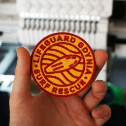 Okrągła, haftowana naszywka z napisem 'LIFEGUARD GDYNIA SURF RESCUE' i motywem deski surfingowej, trzymana w dłoni na tle maszyny do szycia przemysłowej.